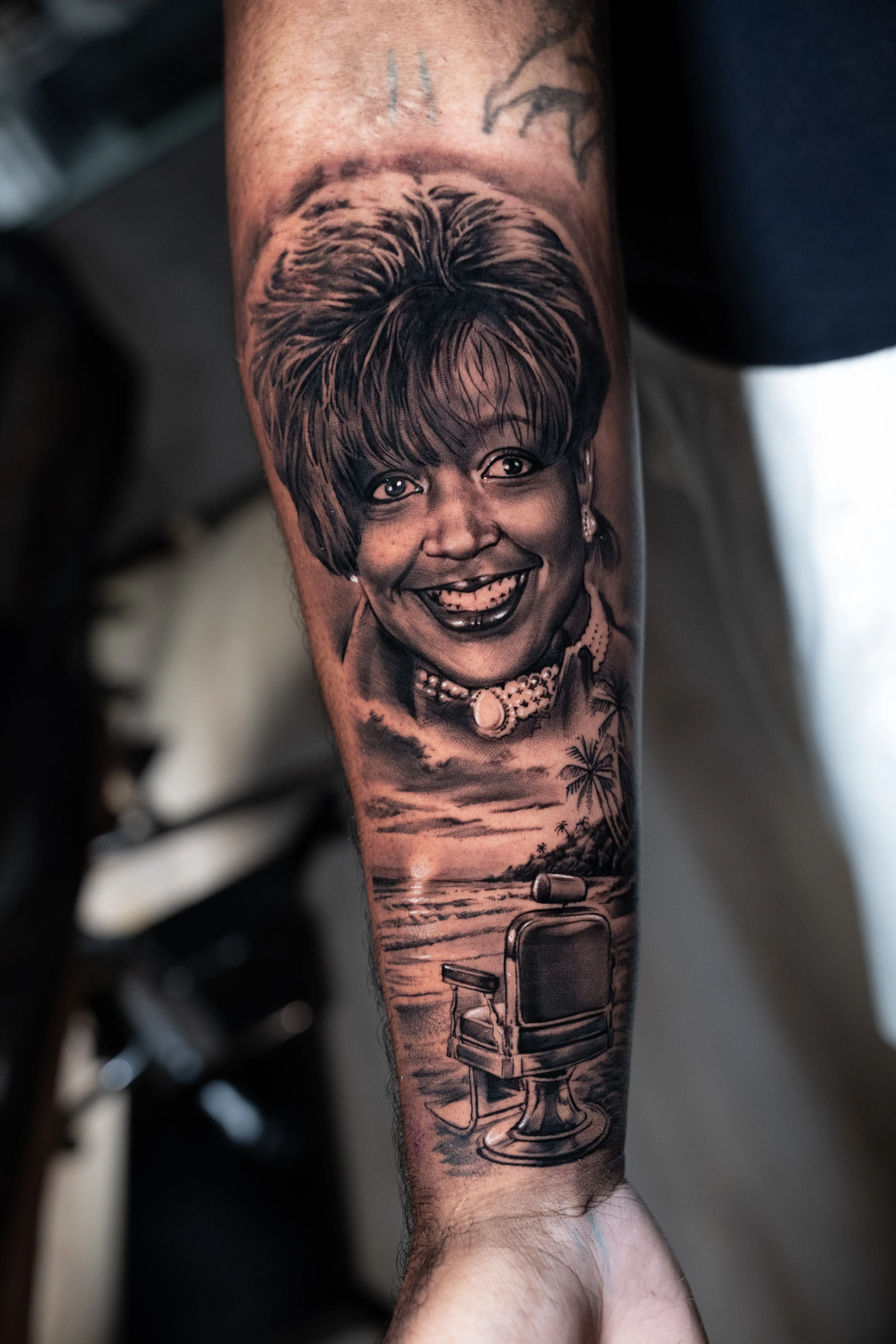 Black-and-grey memorial portrait of a smiling woman with a beach sunset and vintage barber chair — TKOTATTOO