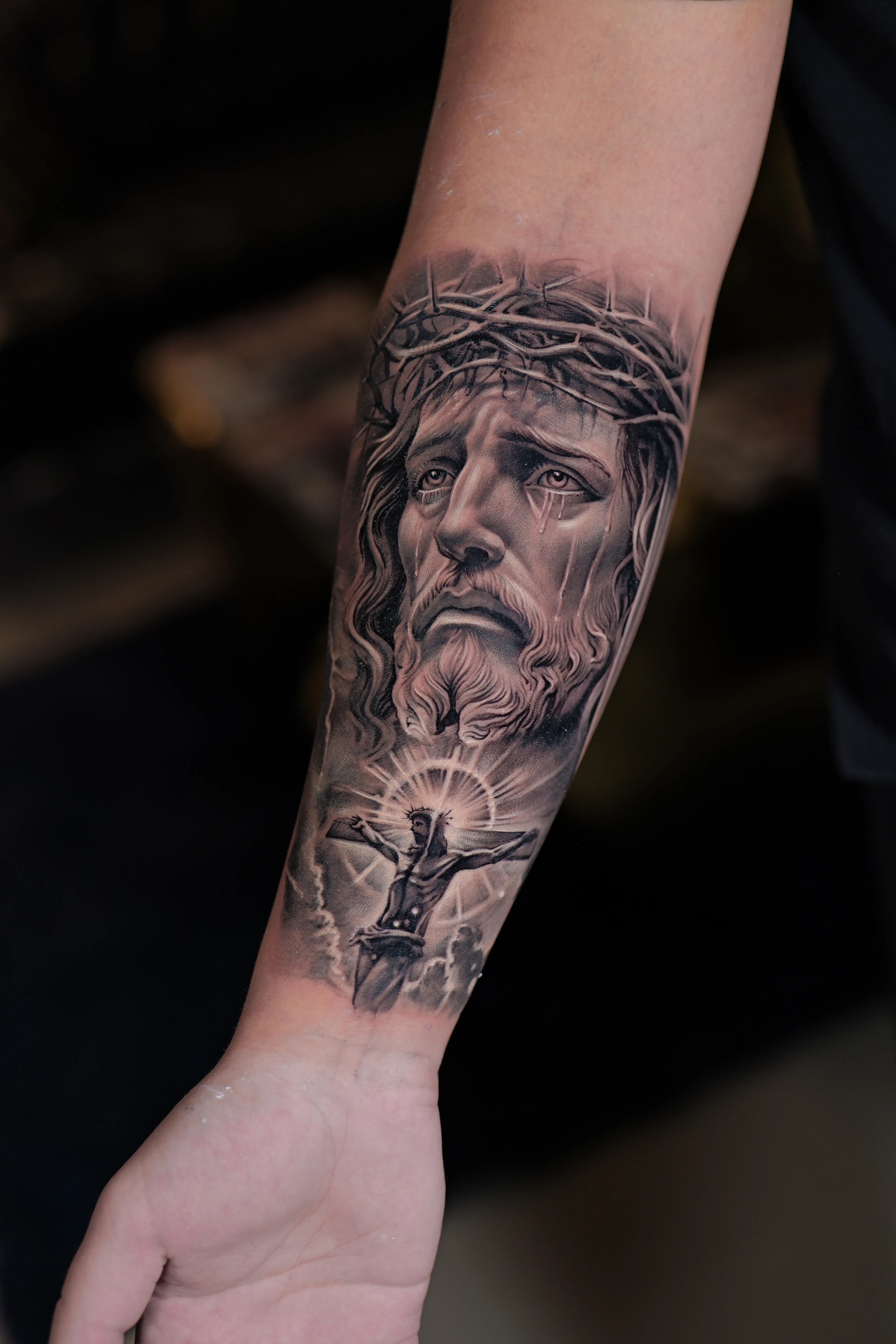 Black-and-grey realism of Christ with the crown of thorns above a glowing crucifixion scene — TKOTATTOO